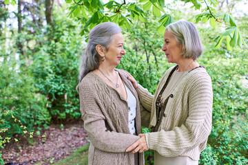 Fototapeta premium A middle-aged lesbian couple in cardigans stand amidst lush greenery, embracing each other with affection.