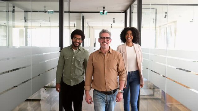 Three colleagues walk confidently down a bright, modern office hallway, smiling as they move towards the camera. Positive and dynamic work environment, emphasizing teamwork and a collaborative spirit