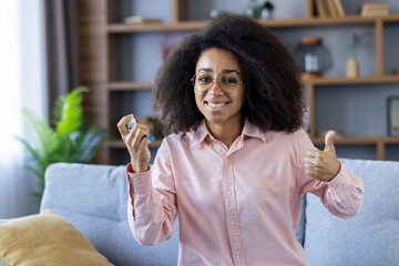 A young woman holding an asthma inhaler, smiling, and showing a thumbs up sign while sitting on a couch at home.