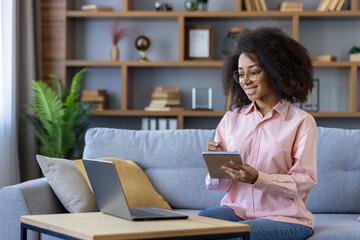 Young student joyfully learning from home, taking notes while engaged in an online video course on laptop. Comfortable home study environment.