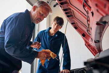 Male Car Auto Mechanic With Trainee Looking Under Bonnet Of Car Checking Oil Level With Dipstick