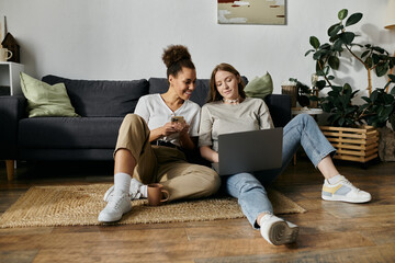 A lesbian couple relaxes at home, one using a laptop and the other looking at a phone.