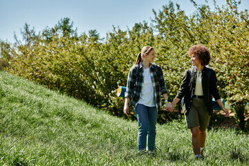 Obraz premium A young, multicultural lesbian couple hikes hand-in-hand through a lush green meadow, enjoying a day outdoors.