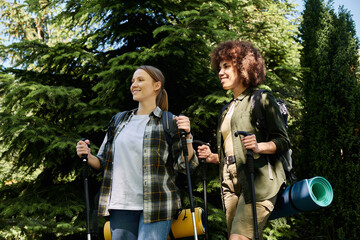 Two young women, dressed in hiking gear, walk through a wooded area on a sunny day.