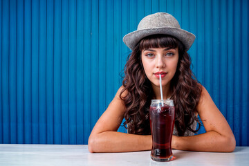 Portrait of a young girl wearing cool hat drinking soda in front a blue blind.	AI Generated