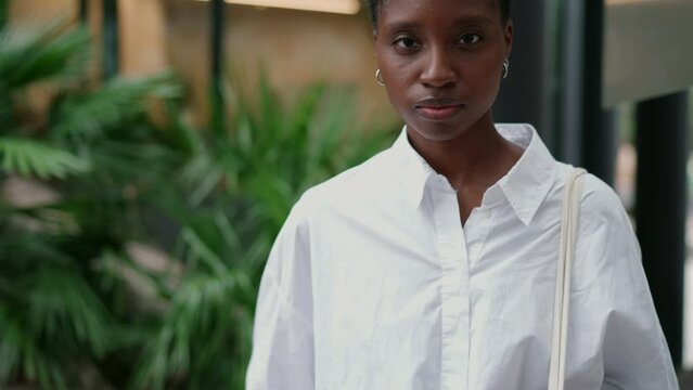 portrait of determined and confident young Black woman standing in modern urban setting posing proudly. Stylish female wearing white shirt and carrying a tote bag, exuding strength and resolve

