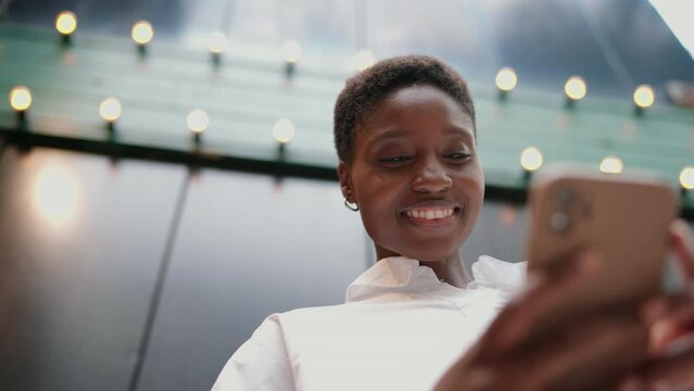 young Black woman laughing joyfully while on phone call. She is standing outdoors against a backdrop of string lights, creating a lively and vibrant atmosphere. Happy female enjoying call on cellular
