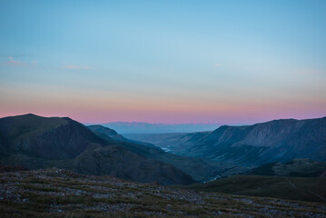 Top view from pass to wide alpine valley with long river in shadow against large mountain range silhouette with snow-capped peaks of vivid pink color far away at sunset. Big snowy ridge on horizon.
