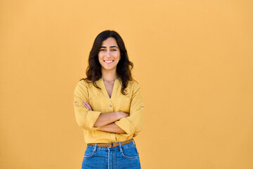 Portrait of middle eastern Israel girl student standing with crossed arms isolated on yellow background. Smiling young Arabian Indian woman, businesswoman, business lady looking at camera, copy space
