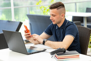 Young male entrepreneur reads a message on his phone and works at his laptop. Work as a freelancer.