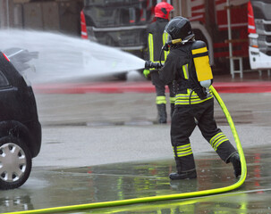 Obraz premium Firefighter with oxygen tank and protective helmet sprays a lot of foam jet on the crashed car