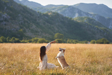 A girl in a flowing dress plays with a joyful Welsh Corgi in a vast meadow, mountains in the...
