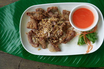Deep-fried pork intestines sprinkled with fried garlic, fried kaffir lime leaves and fried dried chilies and spicy dipping sauce in a white plate placed on a banana leaf.