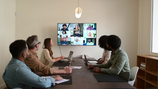 Team engaged in a virtual meeting with a display screen, showing both remote participants and in-person colleagues interacting seamlessly, integration of modern technology in collaborative workspaces
