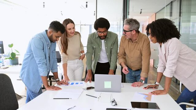 Diverse colleagues gather around a laptop with bright expressions, indicating a successful collaborative effort and a lively brainstorming session in a well-lit, modern workspace, team work concept