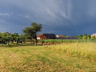 A cargo cloud is visible above the village. a beautiful landscape of fields with yellow grass and a house equipped with solar panels during a thunderstorm. The topic of changes in temperature phenomen