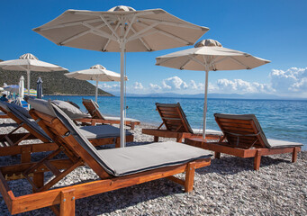 White umbrellas, sunbeds on sandy pebble beach with turquoise crystal clear water in summer morning.