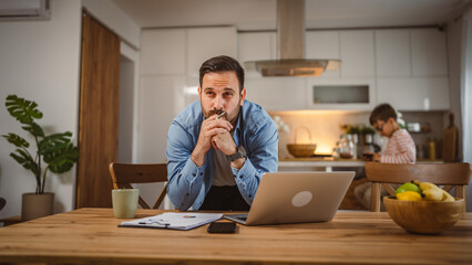 Exhausted man father work at home on laptop with son in the background