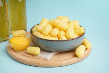 Bowl with tasty salty corn sticks and sauce on blue background, closeup