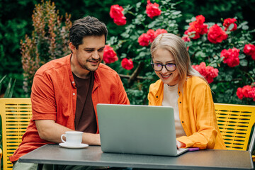 man and woman sitting together at outdoor table in a garden setting with roses, surrounded by vibrant red flowers and lush greenery. Both are engaged with a laptop