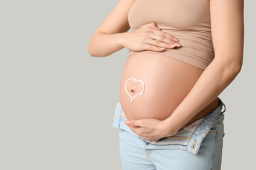 Young pregnant woman with heart made of cream on her belly against grey background