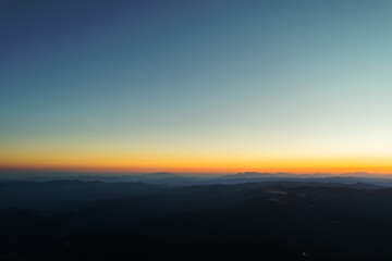 Sunset cloudy sky evening horizon from aerial view. Lens flare in mountains at orange sunset in summer morning aerial view drone. Sun rays. Green pine forest slopes of mountain range. Bright disk sun