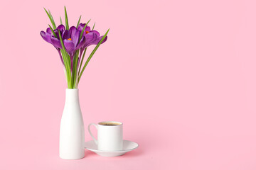 Vase with beautiful crocus flowers and cup of coffee on pink background