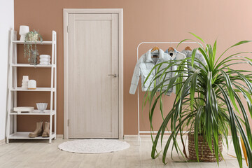 Houseplants, shelving unit and rack with stylish clothes near beige wall in interior of modern...