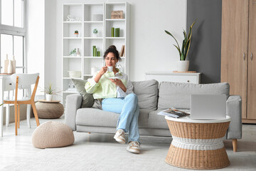 Young African-American woman drinking coffee on sofa at home - Powered by Adobe