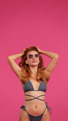 Obraz premium Woman in a stylish grey bikini enjoying the summer vibes with sunglasses on and hands in her hair against a vibrant pink background. Perfect for summer, beach, and fashion themes.