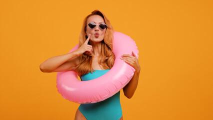 A joyful woman in a turquoise swimsuit with a pink inflatable ring blows a kiss while wearing stylish sunglasses against an orange background. Perfect for summer, vacation, and fun themes.
