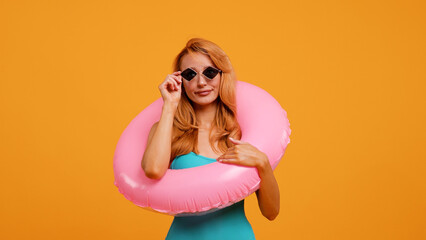 A joyful woman in a turquoise swimsuit with a pink inflatable ring blows a kiss while wearing stylish sunglasses against an orange background. Perfect for summer, vacation, and fun themes.
