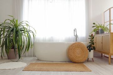 Interior of stylish bathroom with bathtub, chest of drawers, houseplants and window