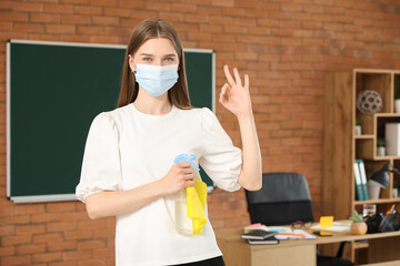 Female teacher with medical mask and detergent showing OK in classroom
