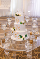 White three-tiered wedding cake with gold petals and white roses. Wedding cake with letters Mr and Mrs on top decorated with gold details on glass round table with candles around