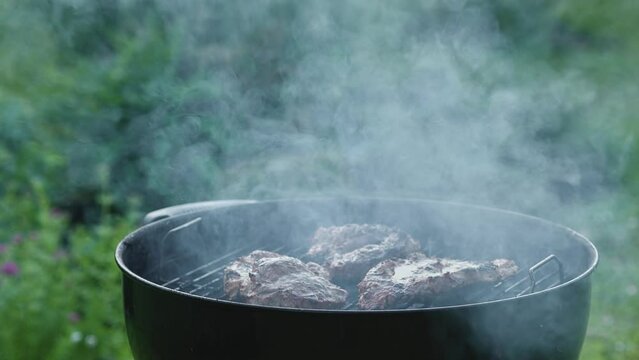 The meat is grilled in the garden. The cook lifts the lid of the grill, smoke is billowing.