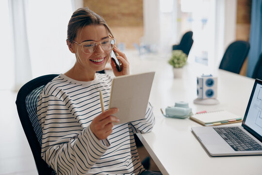 Young female architect talking by phone with client sitting in modern coworking and making notes