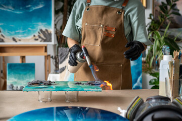 Hands of female artist burn epoxy resin painting with blowtorch, removing air bubbles. Professional...