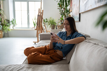 Calm woman with phone relaxing on sofa in home art studio. Resting girl typing message, scrolling through feed on social networks, watching short videos, chatting online on Internet, procrastinating