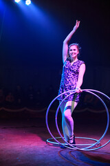 Young slim attractive circus gymnast woman performing live show with hula hoops on red arena with blue backlight, audience is excited