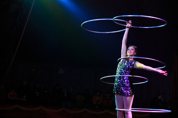 Young slim attractive circus gymnast woman performing live show with hula hoops on red arena with blue backlight, audience is excited
