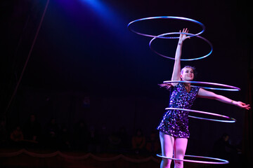 Young slim attractive circus gymnast woman performing live show with hula hoops on red arena with blue backlight, audience is excited