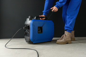 A woman in a blue suit starts a generator. A modern portable gasoline inverter generator stands in a garage against a black wall. Backup autonomous power supply at home. Electricity production.
