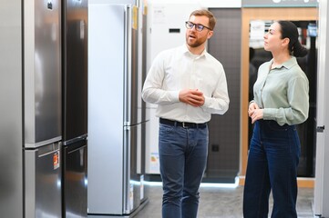 shop assistant demonstrates refrigerator to young woman in home appliance shopping mall supermarket
