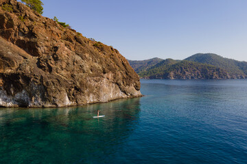 Young woman paddle on sup board at calm sea along rocky coastline. Female doing paddleboarding...