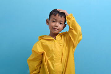Cute little Asian boy wearing a yellow raincoat isolated on blue background. Weather forecast concept