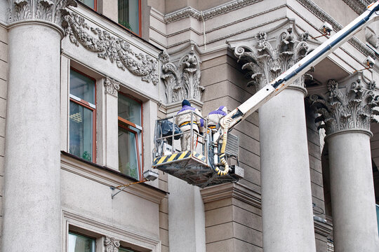Utility workers on lift platform, facade restoration on historic building with ornate columns, preservation of urban architecture. Men on lift platform clean and maintain exterior of historic building