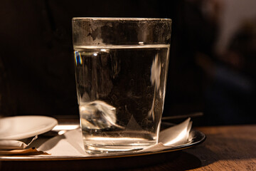 Close up of glass with hot water served on the white plate with napkin, sugar pack and tee bag on the wooden table in Cafe. Dark background. Drink concept. Tea cup. Selective focus. Copy space