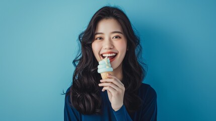 Asian woman with long black hair eating blue ice cream against blue background.