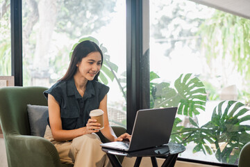 A woman is sitting in a chair with a laptop open in front of her. She is holding a cup of coffee...